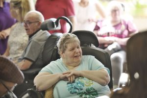 A woman wearing a light blue sequined t-shirt sits in a wheelchair with her hands clasped, smiling and engaged in conversation. Several other people in wheelchairs and staff members are visible in the busy, lively background