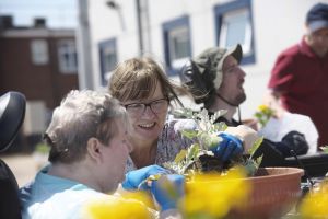 A smiling woman wearing glasses and a floral top leans in closely to assist another person in planting flowers in a terracotta pot outdoors