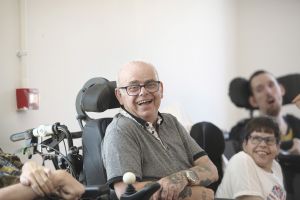 A laughing older man wearing glasses and a grey polo shirt sits in a powered wheelchair with his arms crossed, showing tattoos on his forearms. Two other people in wheelchairs are smiling